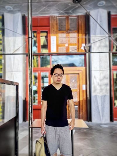 Portrait of young asian man in eyeglasses standing outside storefront under mirrored reflection.