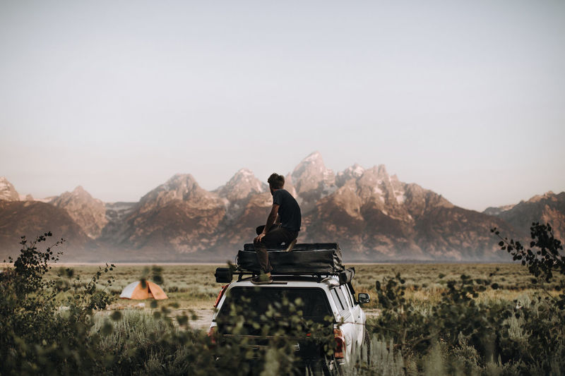 Side view of man sitting on car roof against clear sky in grand teton national park