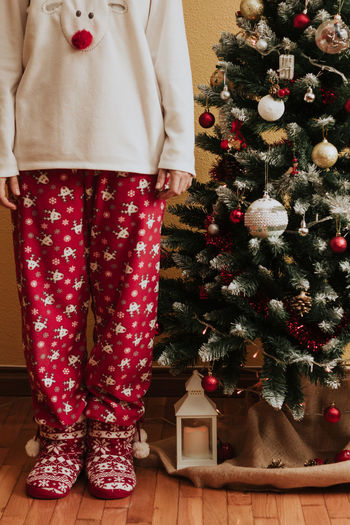 Female in warm pajama standing near decorated spruce on christmas day