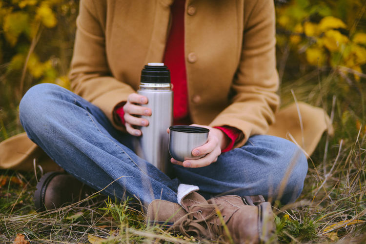 Low section of woman holding insulated drink container while sitting on field during autumn