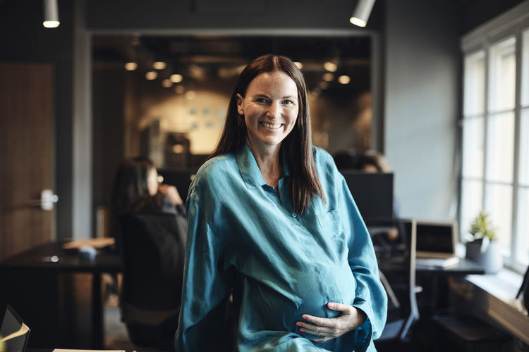 Smiling pregnant female entrepreneur with hand on stomach at office