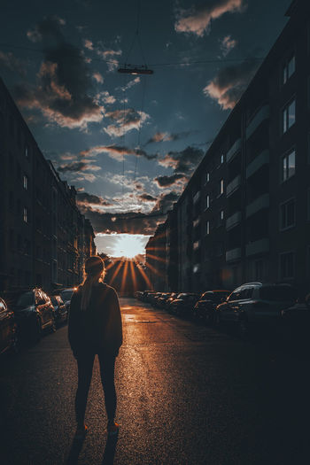 Woman stands on street amidst buildings against sky during sunset