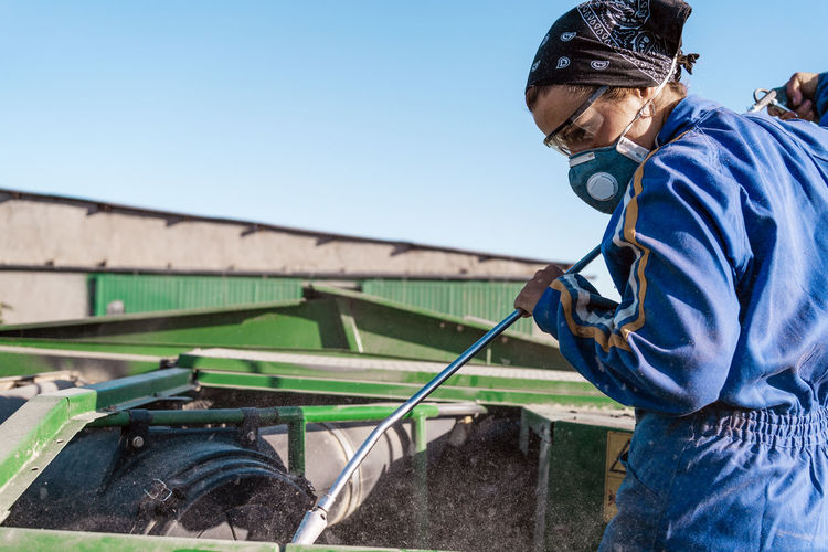 Employee in workwear and protective respirator cleaning agricultural machine in countryside on sunny day