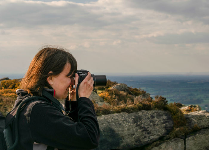 Side view of woman photographing with camera while standing on mountain against cloudy sky during sunset
