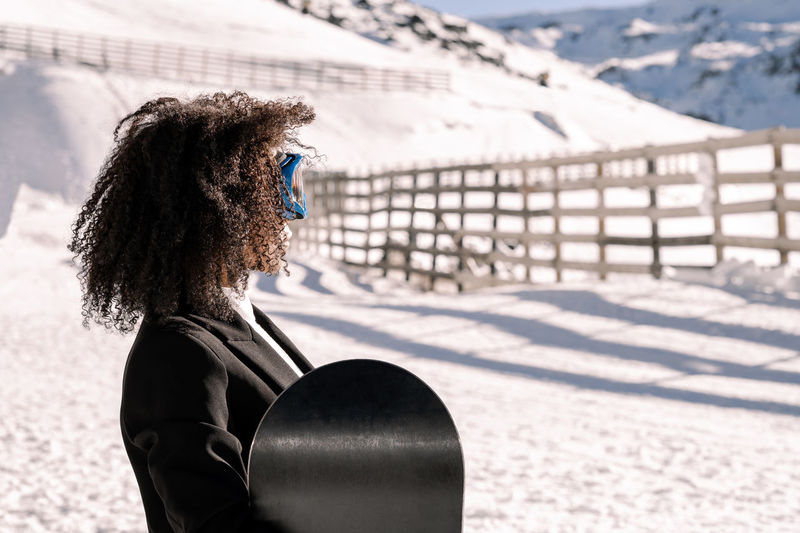 African american woman with goggles and a snowboard on a snowy mountain during winter