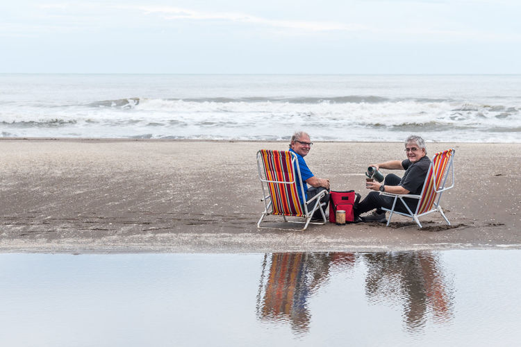 Two mature gray haired men sitting on beach chairs pouring water into a mate, smiling at the camera.