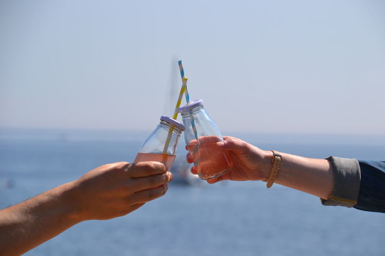 Cropped image of friends hands toasting drinks by sea against sky