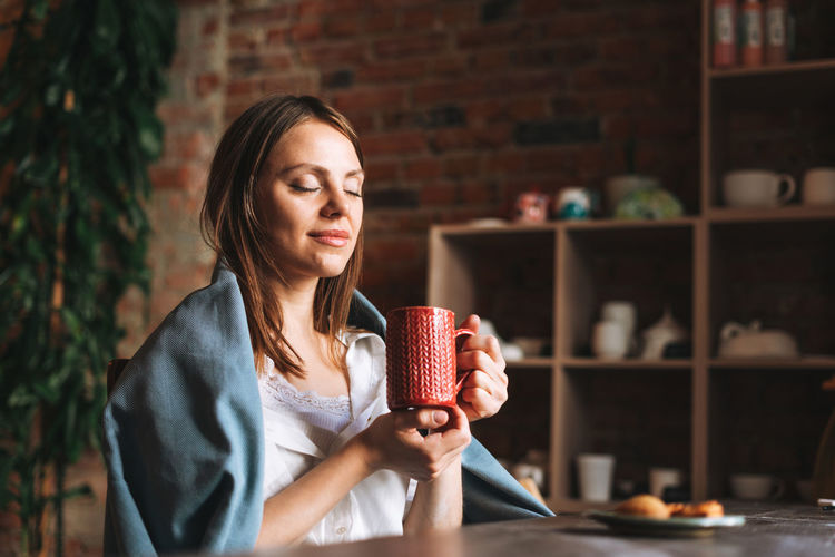 Young woman in cozy grey scarf with mug of tea in hands looks out the window and rests in studio