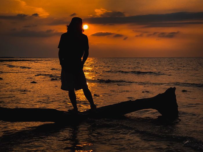 Full length of man standing on driftwood at beach against sky during sunset