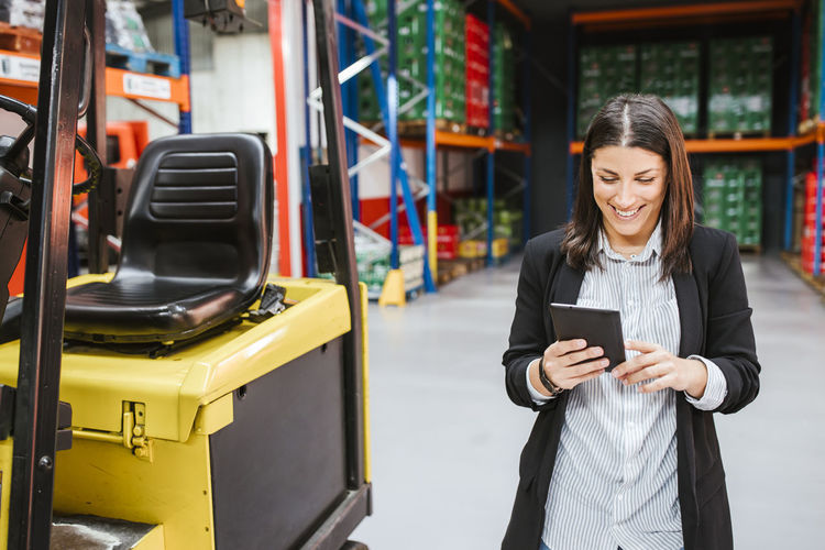 Smiling businesswoman using digital tablet by forklift in distribution warehouse