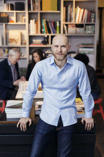 Portrait of confident businessman leaning on desk while colleagues working in background