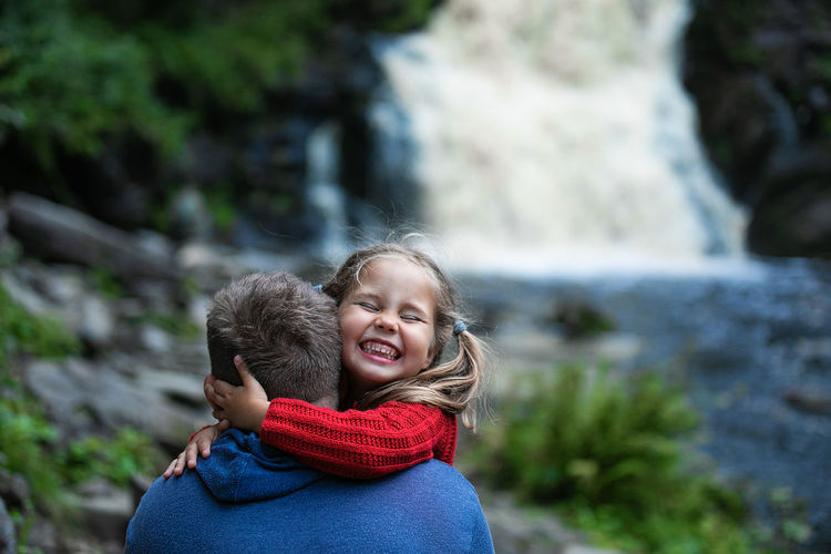 Father carrying smiling daughter against waterfall
