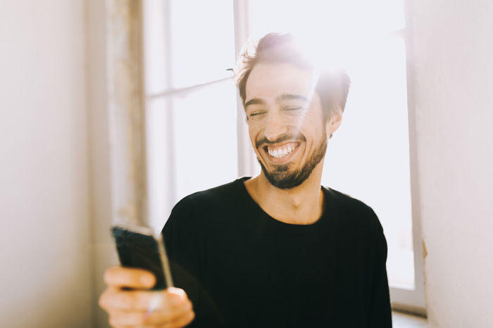 Happy young man using phone by window at home