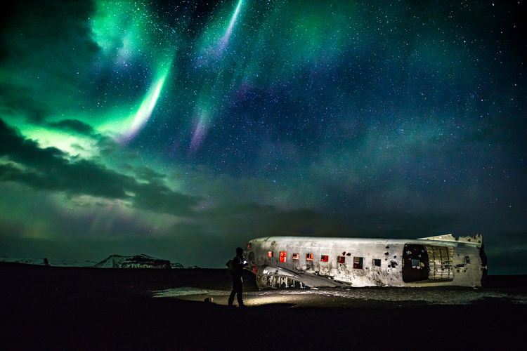 Silhouette hiker by abandoned airplane at beach against aurora borealis during night