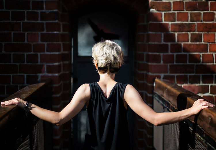 Blond hair woman in a black dress standing with her back on a bridge against with brick wall