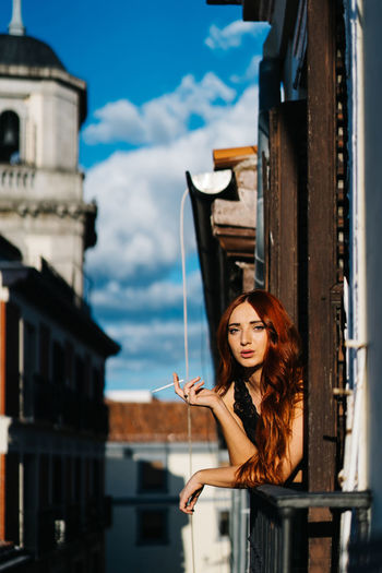 Carefree female with ginger hair leaning on railing on balcony and smoking cigarette while looking at camera in sunny evening