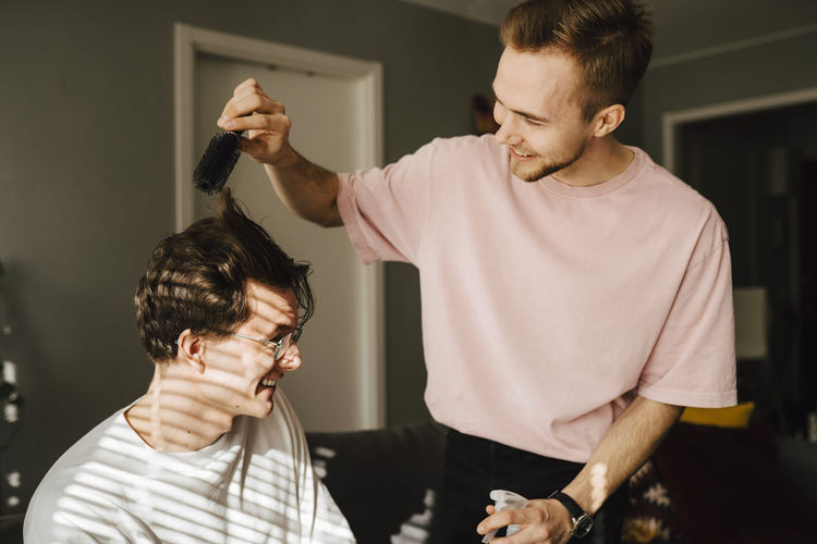Young man combing male friend's hair while sitting at home