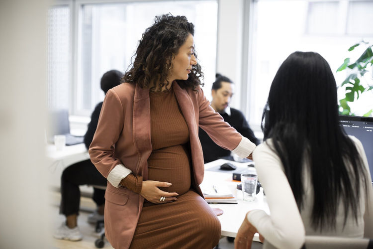 Pregnant businesswoman discussing with female colleague over computer in office
