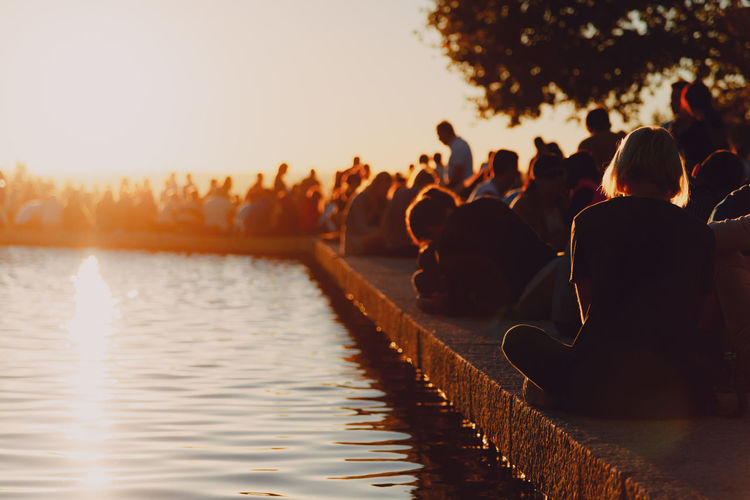 Silhouette people sitting on retaining wall by lake during sunset