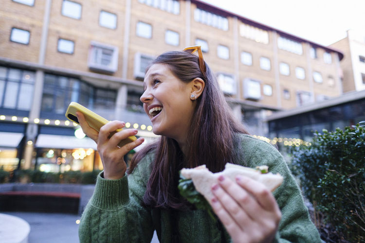 Happy woman holding sandwich talking through speaker of mobile phone in front of building
