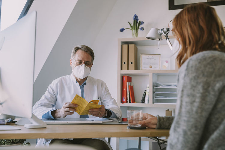 Doctor wearing protective face mask checking patient's vaccination certificate while sitting at office