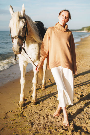 Young woman in beige sweater with white horse on seascape background