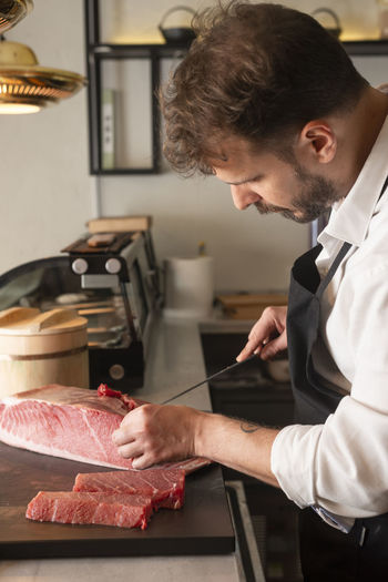 Side view of focused male chef cutting raw fish at table in asian restaurant and preparing sushi