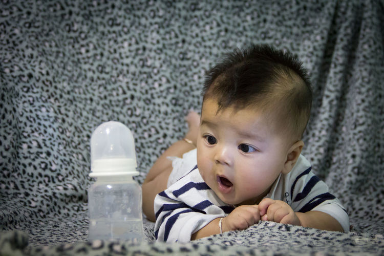 Cute baby boy looking at milk bottle on bed in home