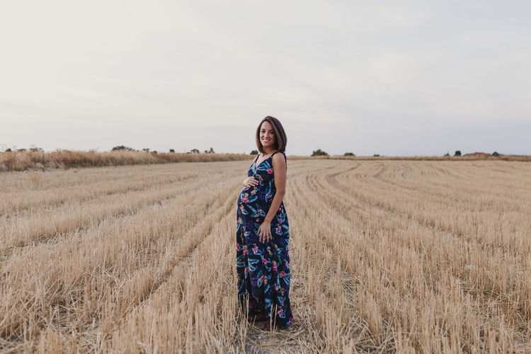 Portrait of smiling pregnant woman with hand on stomach standing on field