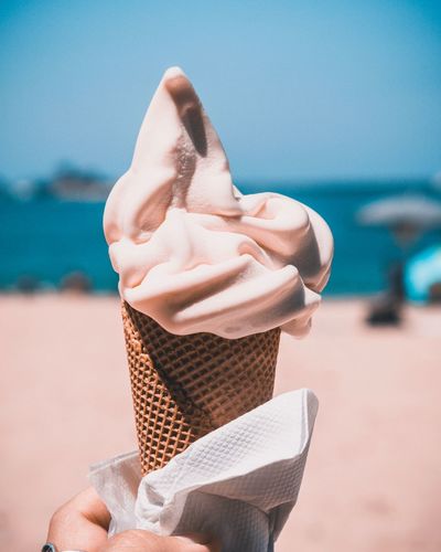 Cropped hand of woman holding ice cream cone at beach against clear blue sky