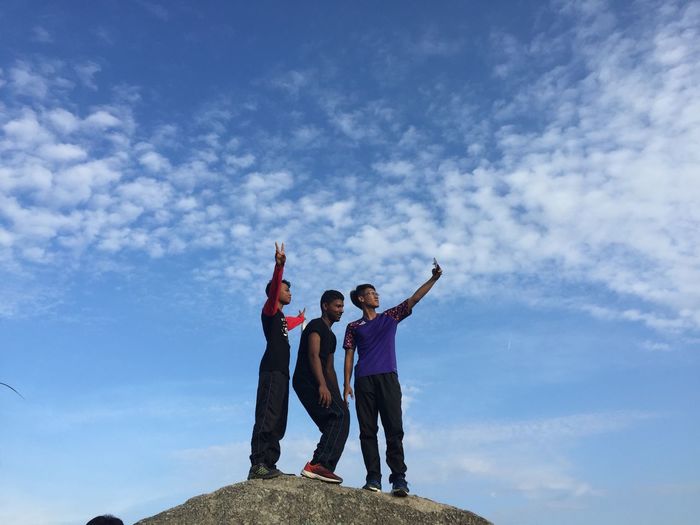 Low angle view of friends with peace sign taking selfie against sky