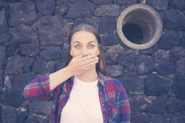Portrait of woman covering mouth while standing against stone wall
