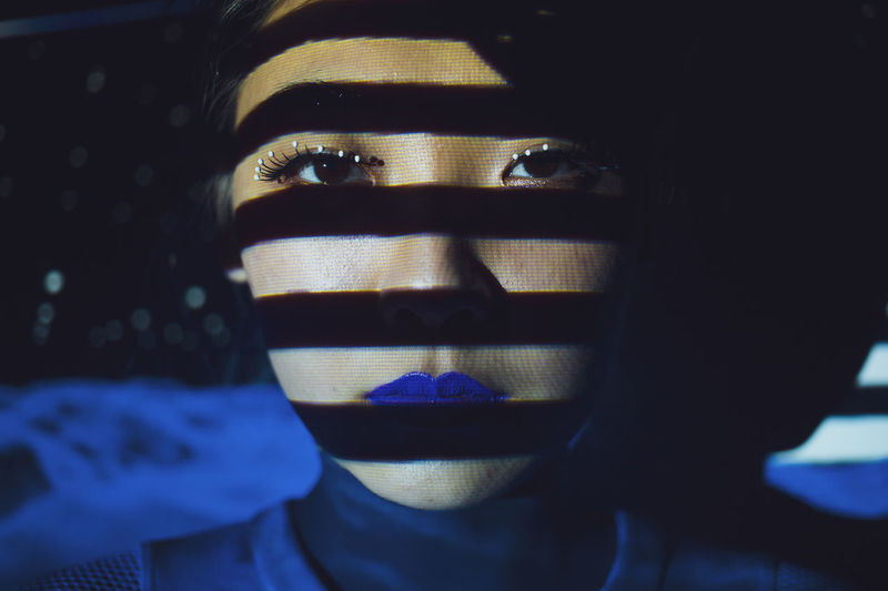 Young asian female model with creative makeup with blue lips and striped shadow on face looking at camera while standing in dark studio with illumination