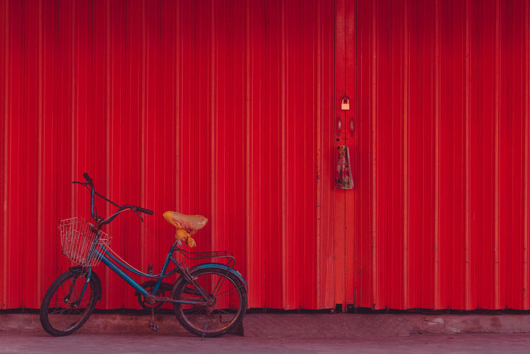 Old weathered blue bicycle parked against red closed shutter door of building.