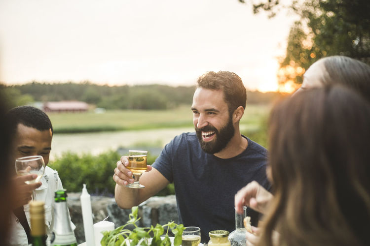Cheerful male and female friends enjoying drink dinner in backyard during sunset