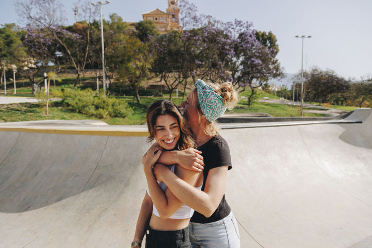 Happy woman embracing friend from behind at skateboard park on sunny day