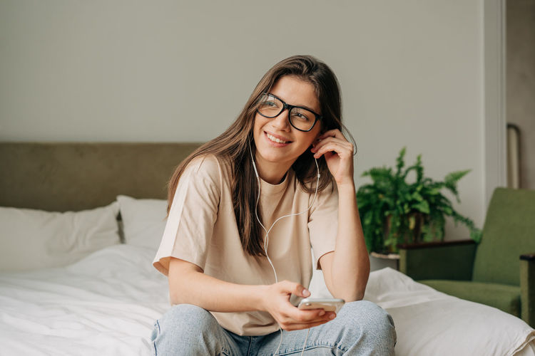 Young happy smiling woman sitting at home listening to music on phone headphones