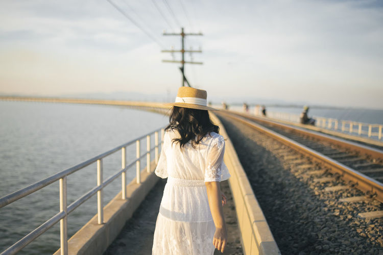 Rear view of woman standing on railroad track by sea against sky