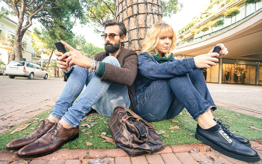Man and woman using mobile phone while sitting against tree trunk in city