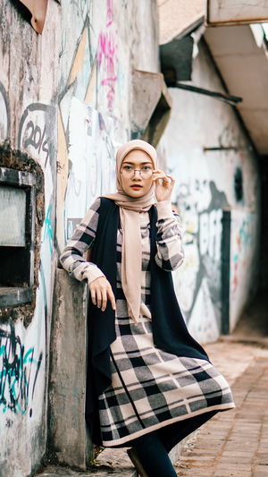 Portrait of young woman wearing hijab and eyeglasses standing against graffiti wall