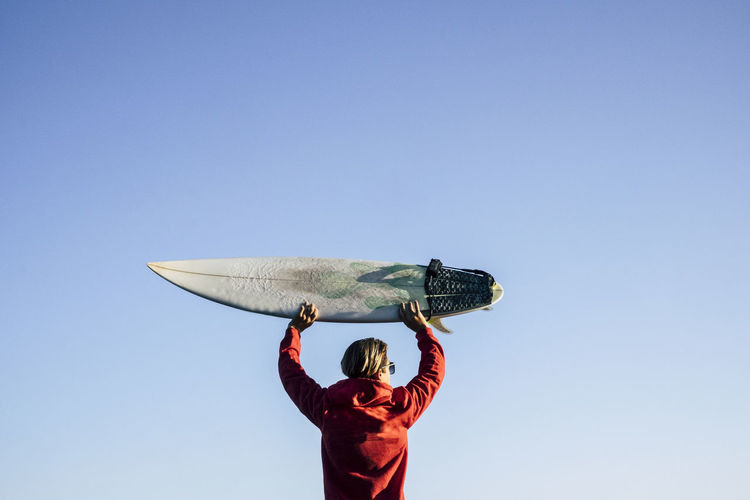 Rear view of teenage boy holding surfboard against clear blue sky