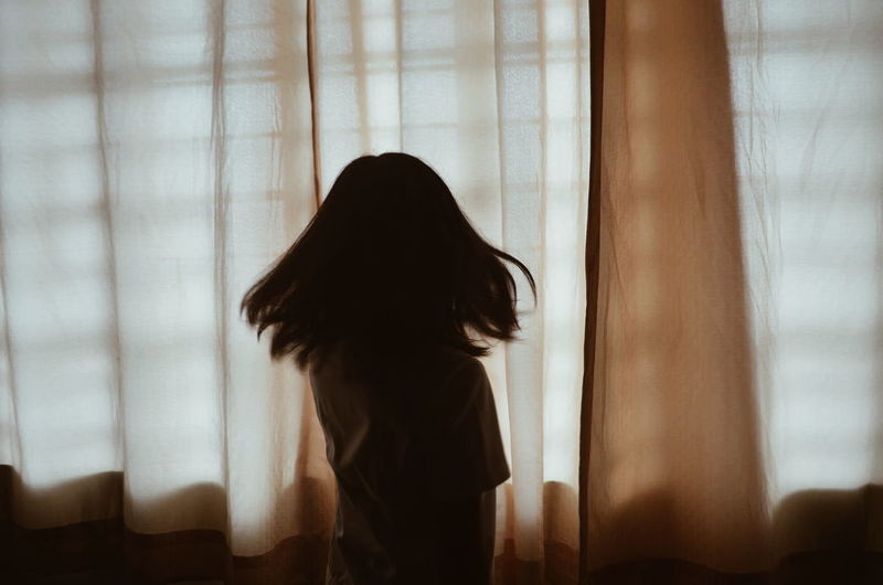 Rear view of girl shaking head by curtain at home