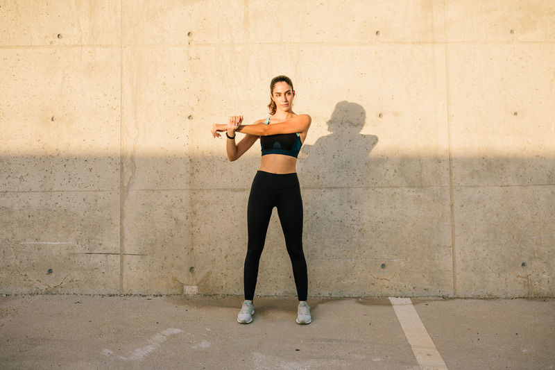 Full body of active young fit female in black sports top and leggings stretching arms while standing against concrete wall in sunny day