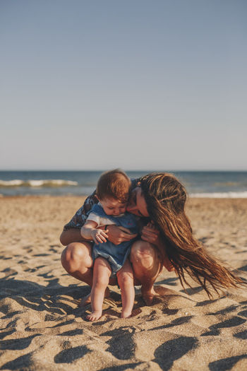 Woman sitting with baby at beach against clear sky