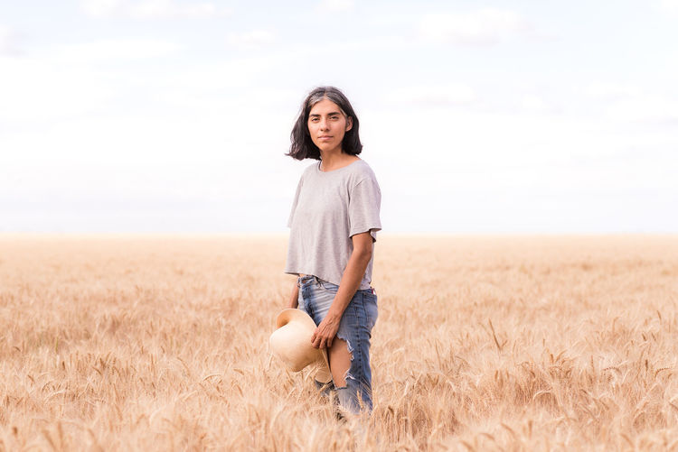 Girl in a wheat field with a hat in her hand and looking at the camera.