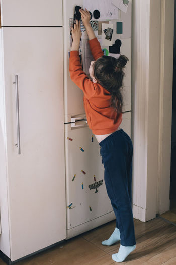 Full length of girl sticking magnet on refrigerator door at home