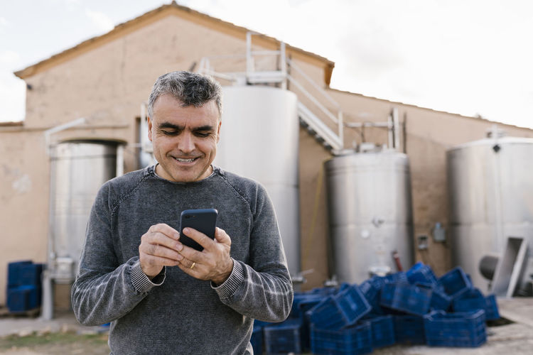 Smiling man using smart phone while standing against wine cellar