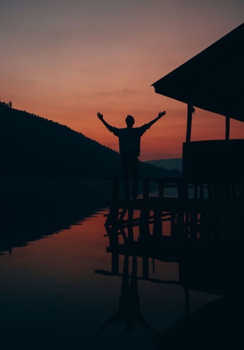 Silhouette man standing while hands raised by lake against sky during sunset