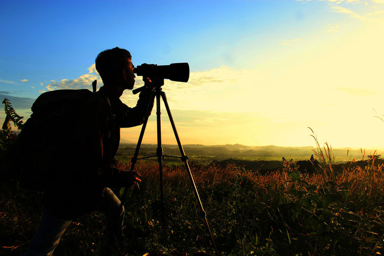 Rear view of man photographing on field against sky during sunset