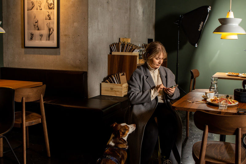 Portrait of young woman sitting on chair in a restaurant with her  dog, looking in her smartphone 